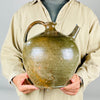 A nineteenth century French terracotta vase with a full rounded body, short flared neck, broad loop handle and separate upward spout. Its glossy green glaze varies through olive and brown, with scattered chips and worn patches revealing the clay beneath. The substantial pottery jug form makes an expressive decorative piece for French country decor.