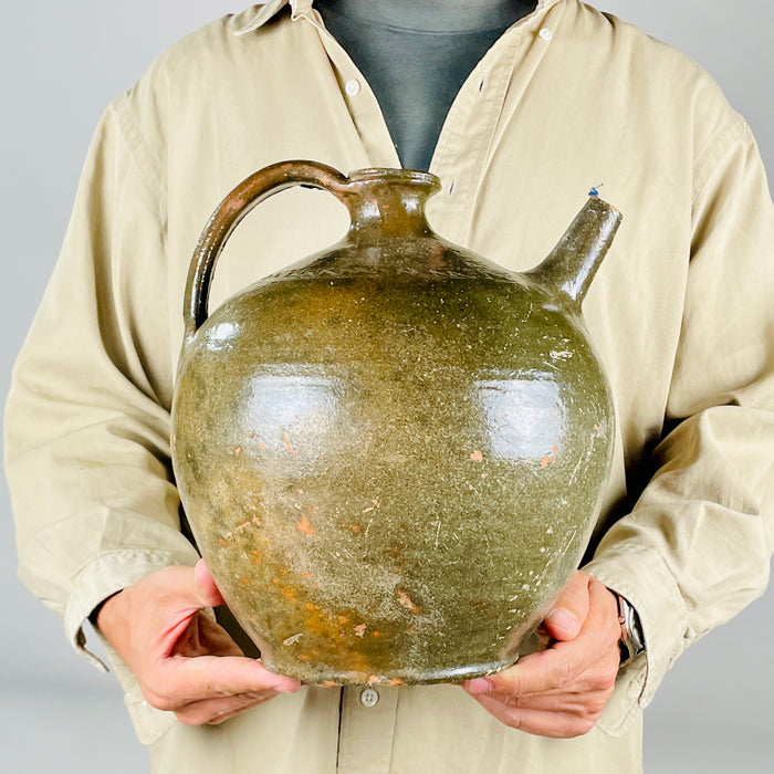 A nineteenth century French terracotta vase with a full rounded body, short flared neck, broad loop handle and separate upward spout. Its glossy green glaze varies through olive and brown, with scattered chips and worn patches revealing the clay beneath. The substantial pottery jug form makes an expressive decorative piece for French country decor.