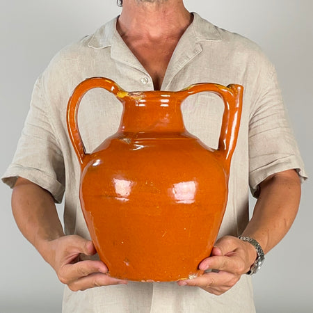 Antique French folk art jug with an open filling mouth and hollow tubular spout, identifying its traditional function as a gargoulette water vessel. Wheel thrown in terracotta during the early 1900s, its glossy brown glaze, exposed red clay and handmade asymmetry give it strong vernacular pottery character.