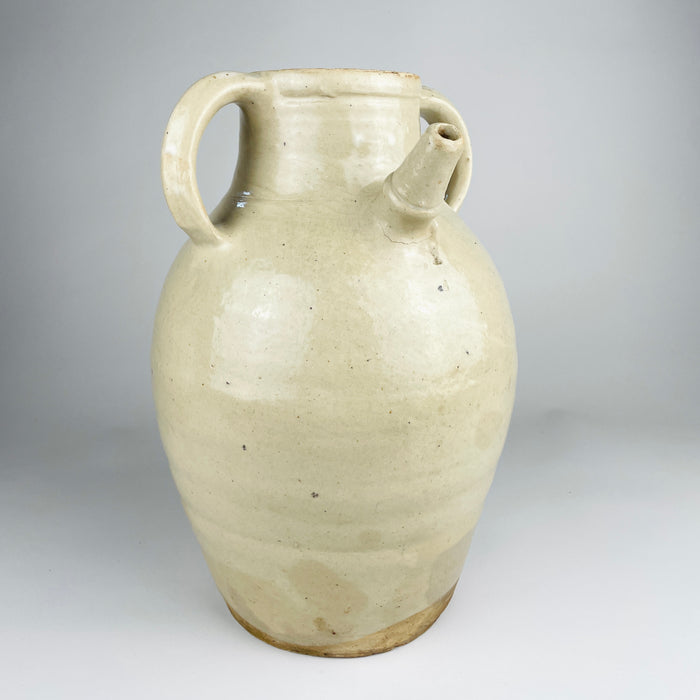 Large antique French white cruche pot from the early 20th century, wheel thrown in glazed terracotta with a rounded body, broad neck, twin loop handles and an applied pouring spout. The cream and ivory surface shows dark mineral specks, age variation and an unglazed foot; the decorative vessel measures 32 cm tall.