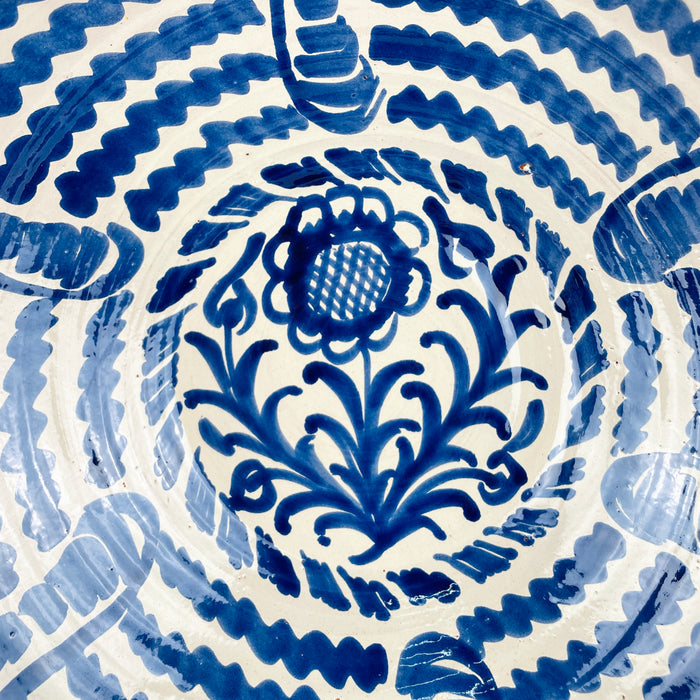 Granada pottery lebrillo bowl with a cream glazed surface and hand painted cobalt blue motifs inside and outside. The interior centers on a sunflower with leafy scrolls and radiating wave bands, while the exterior carries loose elongated blue brushstrokes that echo the handmade decoration. This large Spanish bowl works as wall art, a fruit bowl, or a statement centerpiece for vintage home decor and Spanish decor.