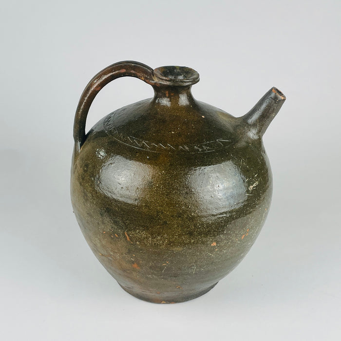 A nineteenth century French terracotta vase with a full rounded body, short flared neck, broad loop handle and separate upward spout. Its glossy green glaze varies through olive and brown, with scattered chips and worn patches revealing the clay beneath. The substantial pottery jug form makes an expressive decorative piece for French country decor.