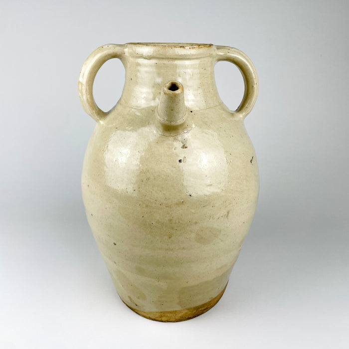 Antique French cruche pot displaying the applied pouring spout, twin loop handles and broad neck that distinguish this regional household vessel. An old hairline follows part of the spout attachment with localized glaze loss, while the surrounding cream terracotta surface retains natural throwing marks and patina.