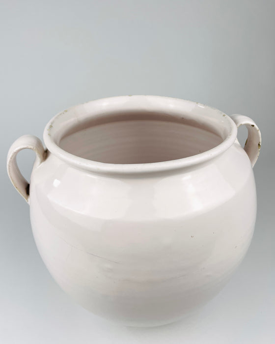 Antique French white confit pot in glazed faience from Martres Tolosane, with a thick curved handle applied to the shoulder and a softly flared rolled lip above. Small glaze chips expose the warm buff ceramic body beneath, adding honest age character to a substantial regional pot à graisse for farmhouse decor.