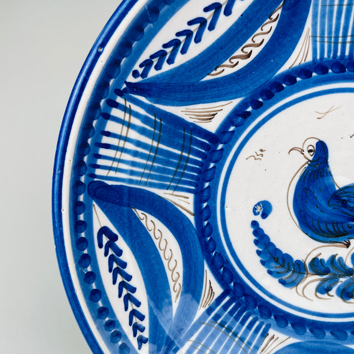 Broad decorative border on the antique Spanish charger combines sweeping cobalt leaves, small blue botanical sprigs, striped lattice sections, dotted rings, and delicate brown wave motifs. These rhythmic hand painted elements surround the central bird and give the Talavera majolica plate the graphic presence associated with collectible European country decor.