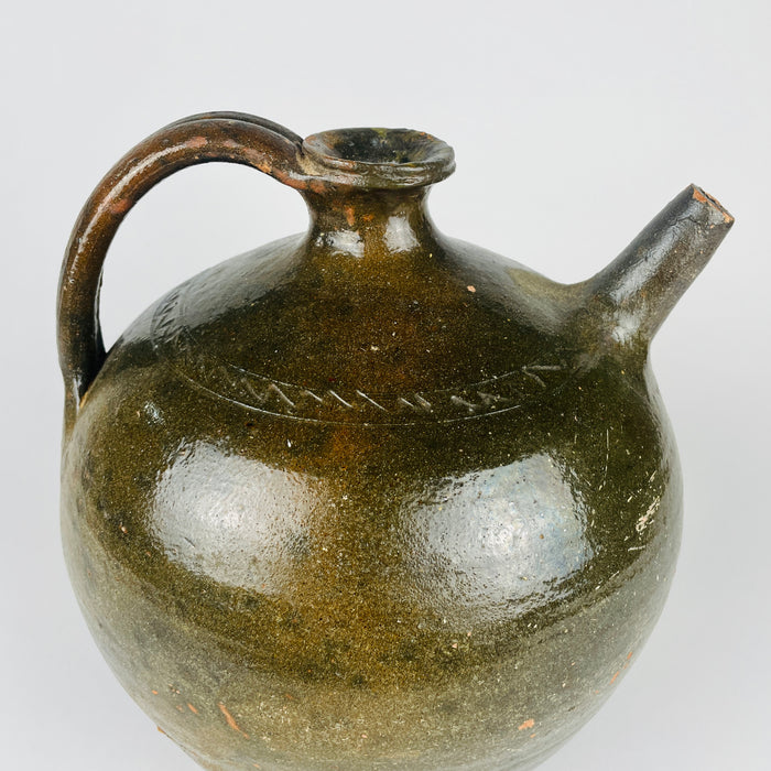 A curved loop handle meets the short flared neck of an antique French terracotta jug. Green and brown glaze covers the handle and rounded shoulder, where an incised zigzag border adds decoration. Worn edges and scattered exposed clay give this traditional pottery vase its individual surface character and emphasise the handmade construction.