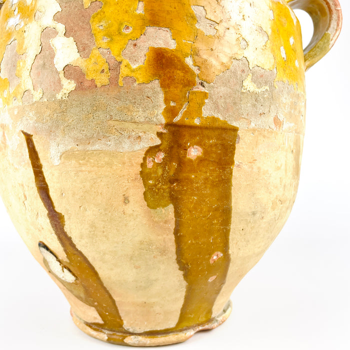 Antique French confit pot in pottery and terracotta, with yellow glaze, rolled rim, and distinctive hand-applied handle detail. The surface cararies heavy patina, glaze crazing, and worn patches that emphasize age and handmade construction. This organic decorative vessel suits wabi sabi decor, French country decor, and rustic interiors where weathered materials and antique pottery are central.