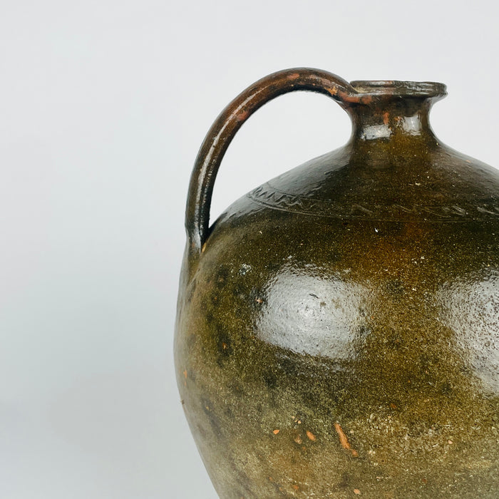 An antique French pottery jug has a glossy dark green body with a broad patch of missing glaze, exposing warm orange terracotta beneath. Its arched handle rises beside the short neck, opposite the projecting spout. The contrast between glazed and worn areas gives this decorative terracotta vase rich texture for rustic home decor.