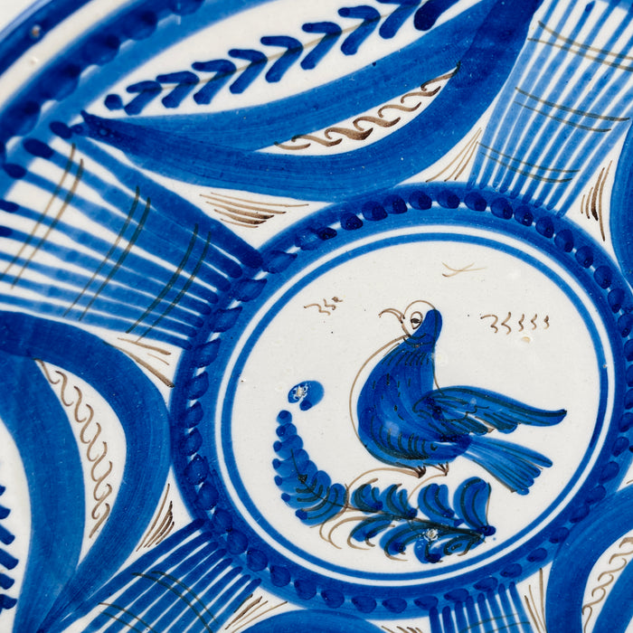 Central decoration on the Spanish Talavera wall plate depicts a stylized cobalt blue bird standing over a plume shaped branch, outlined and feathered with fine manganese brown strokes. The motif sits within double blue rings on a white tin glazed ground, giving this antique faience bird plate a clear focal point for botanical wall art.