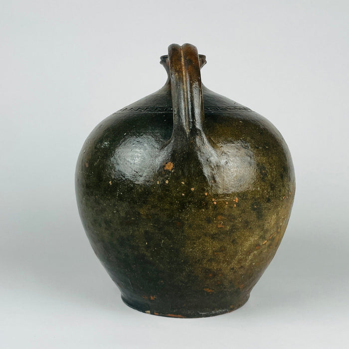 The thick flared rim of an antique French terracotta jug meets a broad curved handle above the rounded body. Olive green and warm brown glaze covers the uneven surface, with worn patches and fine lines visible around the join. These construction and condition details reveal the handmade qualities of this substantial French pottery vessel.