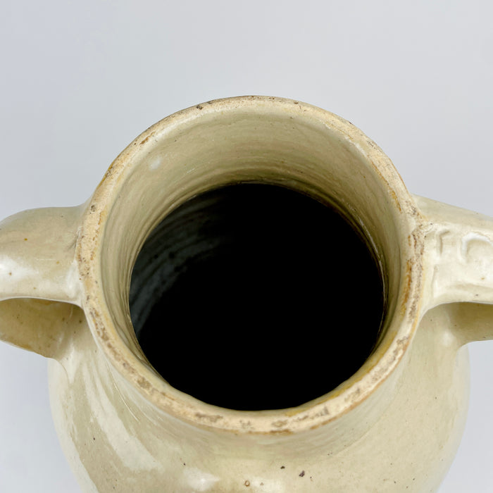Antique white terracotta water jug from France with concentric throwing lines visible through the glossy cream glaze. Small iron-colored specks and tonal patches animate the generous body, while the twin handles and compact pouring spout preserve the recognizable form of a traditional cruche or pottery vase.