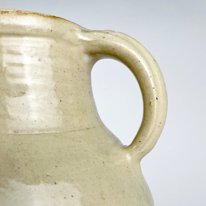 French terracotta water cruche with a narrow applied spout projecting from the upper shoulder between two handles. The cream glaze pools and changes tone around the handmade join, with an old hairline and small glaze losses visible at the attachment; this age detail is part of the antique vessel’s condition.