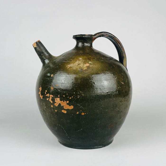 An antique French green glazed terracotta jug has a broad rounded body, short flared neck and arched handle opposite a narrow pouring spout. An incised zigzag band follows the shoulder above the gently tapering base. The olive and brown pottery vessel can also be styled as a decorative vase or table centerpiece within Mediterranean decor.