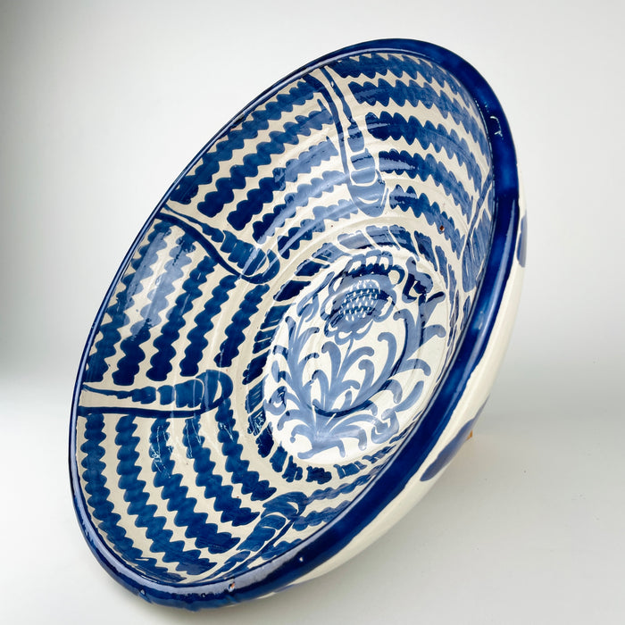 Large Spanish decorative bowl in the Fajalauza tradition, formed from terracotta earthenware and finished with white glaze and cobalt blue hand painting. The broad lebrillo shape frames a sunflower motif, layered foliage, and repeated wavy bands, producing a bold graphic surface. Its generous scale makes it suited to Mediterranean decor, rustic home decor, cottage decor, and a blue and white ceramic collection.