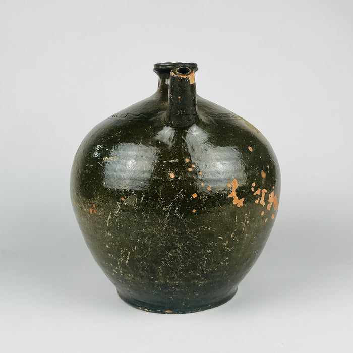 An antique French green glazed terracotta jug has a broad rounded body, short flared neck and arched handle opposite a narrow pouring spout. An incised zigzag band follows the shoulder above the gently tapering base. The olive and brown pottery vessel can also be styled as a decorative vase or table centerpiece within Mediterranean decor.