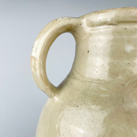 Early 1900s French cruche pot formed on a potter’s wheel from fired clay and covered in an irregular cream glaze. The broad cylindrical neck, symmetrical loop handles and small projecting spout identify its traditional household vessel form, while its generous scale gives this antique pottery strong decorative presence.