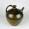 An antique French terracotta jug has an enclosed rounded body, a flared filling neck and a separate narrow spout for pouring. Green glaze varies through olive and brown, with small areas of orange clay showing through wear. Handworked contours and simple incised decoration give this ceramic vessel the distinctive character of traditional French country pottery.