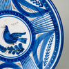 Antique Spanish faience plate with a lively bird, plume shaped branch, curved leaves, botanical sprigs, dotted bands, and gridded panels, all painted by hand over an opaque white glaze. The combination of aged ceramic texture and disciplined cobalt ornament gives this Talavera charger a strong role in rustic home decor.
