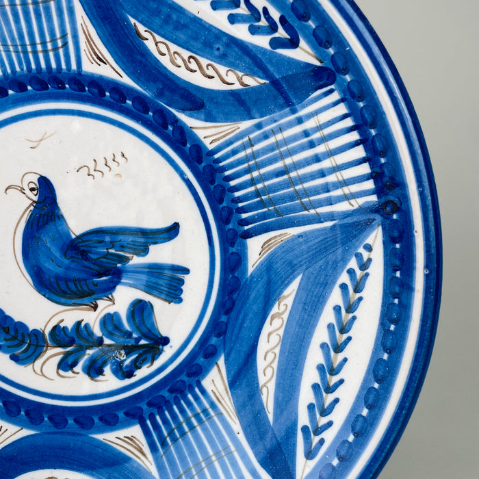 Antique Spanish faience plate with a lively bird, plume shaped branch, curved leaves, botanical sprigs, dotted bands, and gridded panels, all painted by hand over an opaque white glaze. The combination of aged ceramic texture and disciplined cobalt ornament gives this Talavera charger a strong role in rustic home decor.