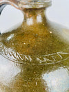 The tubular spout of an antique French pottery pitcher rises from its rounded shoulder, with warm orange clay exposed around the opening. Small glaze losses and pale scratches extend across the broad green glazed body. These visible surface details distinguish the historic pouring vessel and give the terracotta pottery its characteristically worn finish.