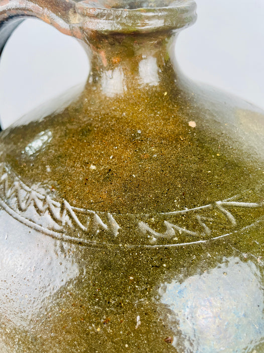 The tubular spout of an antique French pottery pitcher rises from its rounded shoulder, with warm orange clay exposed around the opening. Small glaze losses and pale scratches extend across the broad green glazed body. These visible surface details distinguish the historic pouring vessel and give the terracotta pottery its characteristically worn finish.