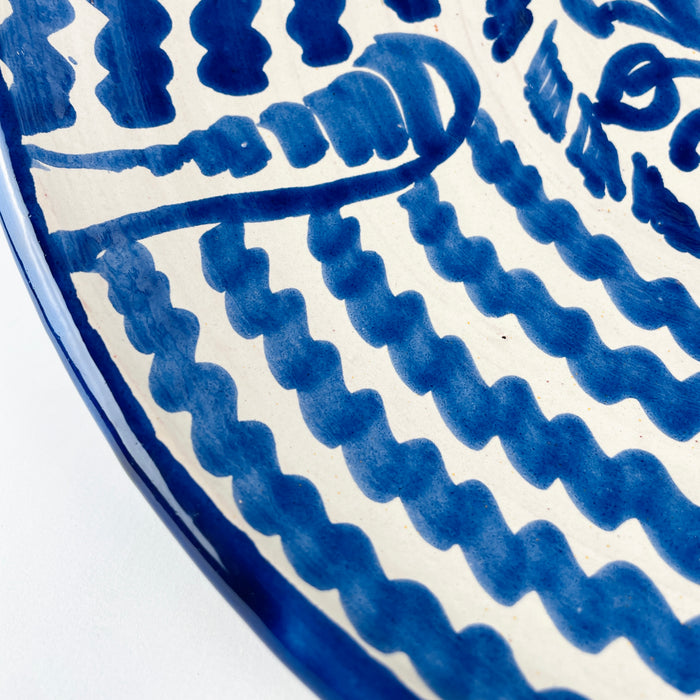 Blue and white Spanish lebrillo bowl from Granada, hand painted with a stylized sunflower and scrolling botanical motifs at the center, surrounded by concentric bands of irregular cobalt waves. The rounded basin, painted rim, and raised foot preserve the traditional form of Fajalauza pottery. It can be used as a decorative wall hanging, fruit bowl, or table centerpiece in farmhouse decor and Mediterranean decor.