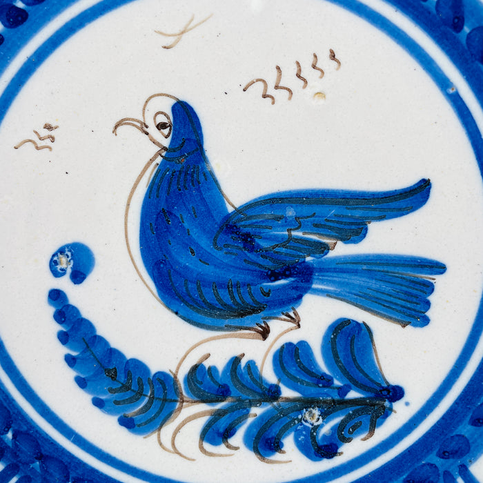 Central decoration on the Spanish Talavera wall plate depicts a stylized cobalt blue bird standing over a plume shaped branch, outlined and feathered with fine manganese brown strokes. The motif sits within double blue rings on a white tin glazed ground, giving this antique faience bird plate a clear focal point for botanical wall art.