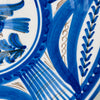 Blue and white Spanish bird charger displayed as a decorative focal object beside glass and ceramic vessels, showing how its circular silhouette and saturated cobalt painting hold their own within a layered arrangement. The early 20th century Talavera wall plate also pairs naturally with coastal decor built around crisp white and marine blue tones.