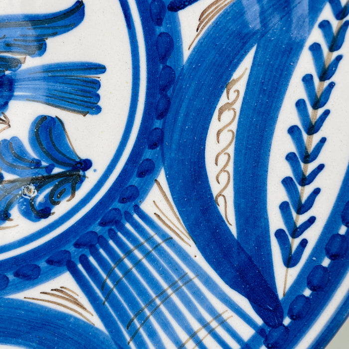 Blue and white Spanish bird charger displayed as a decorative focal object beside glass and ceramic vessels, showing how its circular silhouette and saturated cobalt painting hold their own within a layered arrangement. The early 20th century Talavera wall plate also pairs naturally with coastal decor built around crisp white and marine blue tones.