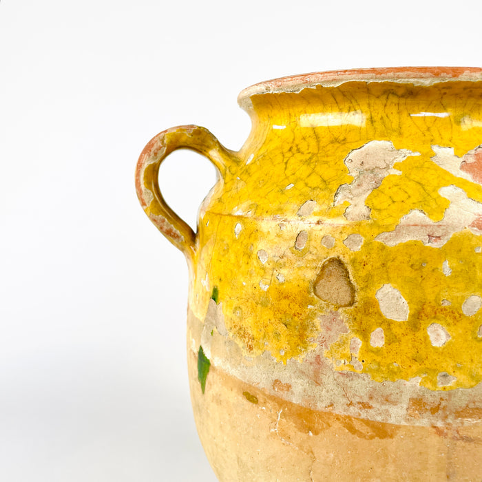 Antique French confit pot in handmade pottery with yellow glaze over the upper body and interior, paired with an unglazed lower half in pale clay. The twin-handle form, rounded profile, and worn terracotta surface identify it as a classic French preserving jar. This decorative antique vessel works as French pottery decor, a rustic vase, or a sculptural accent in country interiors.