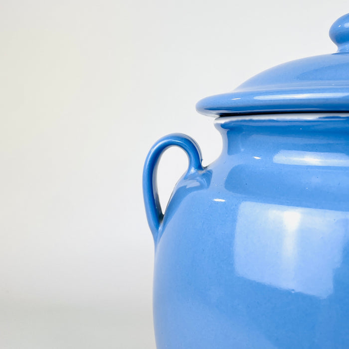 Early 20th century French confit pot is held at torso height to communicate its generous scale and rounded proportions. Its glossy cornflower-blue glaze, matching lid, twin handles and cream interior identify a Martres Tolosane faience storage jar suited to a console, kitchen shelf or decorative pottery collection.