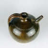 The rounded body of an antique French green glazed terracotta vase has olive, brown and dark moss tones interrupted by scratches and small patches of exposed orange clay. A short neck, curved handle and tubular spout complete the form. Its visibly worn pottery surface and irregular patina complement the natural textures associated with wabi sabi decor.