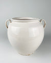 Extra large antique French white confit pot from Martres Tolosane, shown held at torso height to communicate its substantial scale. The early 20th century faience jar has a rounded wheel thrown body, opaque ivory glaze, wide rolled rim and two applied loop handles, making it a sculptural ceramic vessel for a console or large tabletop.