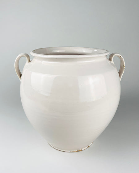 Extra large antique French white confit pot from Martres Tolosane, shown held at torso height to communicate its substantial scale. The early 20th century faience jar has a rounded wheel thrown body, opaque ivory glaze, wide rolled rim and two applied loop handles, making it a sculptural ceramic vessel for a console or large tabletop.