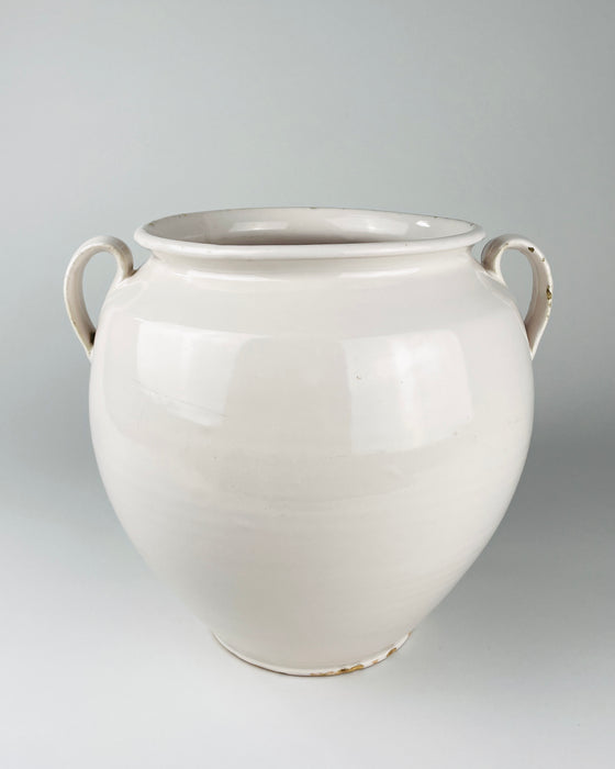 Extra large antique French white confit pot from Martres Tolosane, shown held at torso height to communicate its substantial scale. The early 20th century faience jar has a rounded wheel thrown body, opaque ivory glaze, wide rolled rim and two applied loop handles, making it a sculptural ceramic vessel for a console or large tabletop.