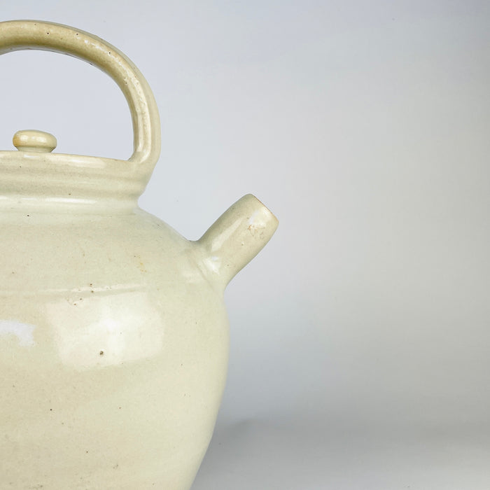 Antique French pottery jug showing one substantial loop handle joined between the rolled rim and rounded shoulder. Variations in the ivory glaze, scattered dark specks and light rubbing reveal the handmade fired-clay surface of this early 20th century cruche pot, now presented as a decorative folk object.