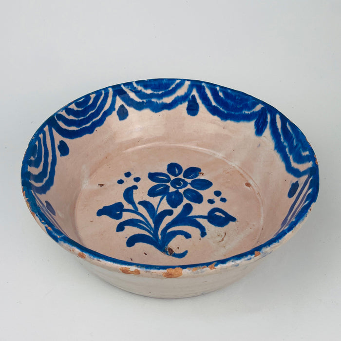 Ceramic bowl with blue floral design on a white background. Spanish terracotta wabi-sabi bowl with opaque tin glaze and cobalt blue floral motif, shaped as a traditional lebrillo serving bowl and displayed as wall hanging ceramic decor using the attached wire on the back. The border shows layered wave brushwork. Notable rim chips and glaze losses are present, consistent with age. Ideal for vintage kitchen styling and Mediterranean decor.