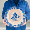 Person holding a Granada Spanish pottery bowl, Fajalauza, painted in cobalt blue with a central flower and surrounding dotted accents, plus a bold wave rim border. Opaque tin glaze over terracotta gives a soft cream tone. Back retains hanging wire for wall mounting. Suitable for cottage decor, farmhouse decor, 