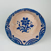 Ceramic plate with blue floral design on a beige background. Cobalt blue tin-glazed Spanish bowl likely associated with Granada Fajalauza pottery, featuring a hand-painted flower in the center and a wave-and-teardrop border. Deep serving profile suits Mediterranean table settings for tapas, fruit, or bread. Reverse shows wire hanger for wall display.