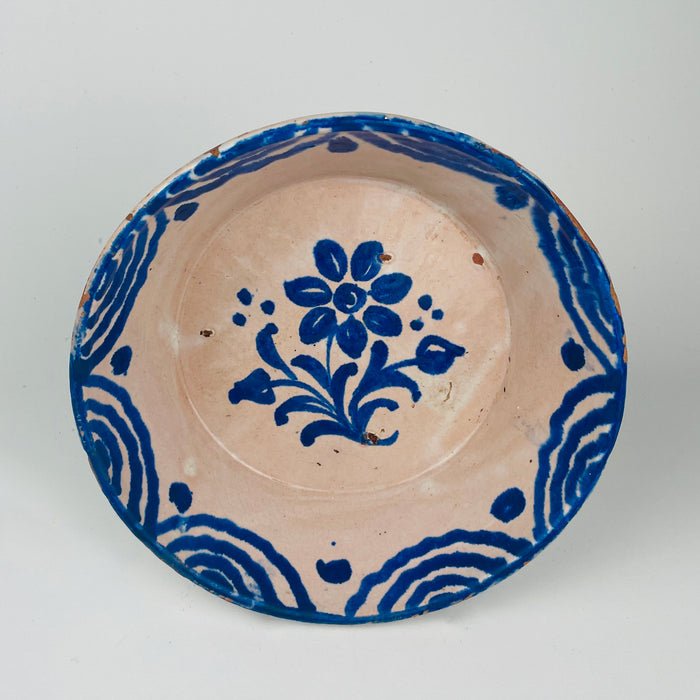 Ceramic plate with blue floral design on a beige background. Cobalt blue tin-glazed Spanish bowl likely associated with Granada Fajalauza pottery, featuring a hand-painted flower in the center and a wave-and-teardrop border. Deep serving profile suits Mediterranean table settings for tapas, fruit, or bread. Reverse shows wire hanger for wall display.