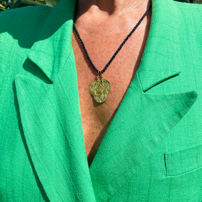 Angled close-up highlighting the hammered coin pendants, each 2 cm wide, reflecting light with a golden shimmer. The artisanal surface texture contrasts with the matte black braided cord, underscoring its role as lightweight minimalist jewelry.