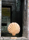 Scenic outdoor view of the hand-carved wooden shell sculpture by ROC Objects, photographed against the backdrop of ancient stone architecture. The acacia wood's warm tones and scallop-inspired fluted ridges contrast beautifully with the mossy green stone textures, highlighting its natural artistry. This image situates the piece within a timeless, earthy environment, reinforcing its alignment with wabi-sabi, organic modern, and contemporary rustic interiors where natural materials and sculptural forms create