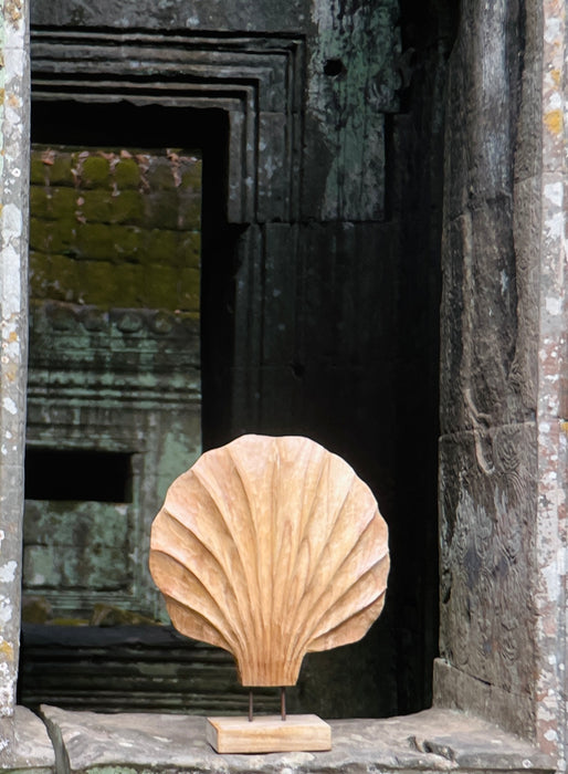 Scenic outdoor view of the hand-carved wooden shell sculpture by ROC Objects, photographed against the backdrop of ancient stone architecture. The acacia wood's warm tones and scallop-inspired fluted ridges contrast beautifully with the mossy green stone textures, highlighting its natural artistry. This image situates the piece within a timeless, earthy environment, reinforcing its alignment with wabi-sabi, organic modern, and contemporary rustic interiors where natural materials and sculptural forms create