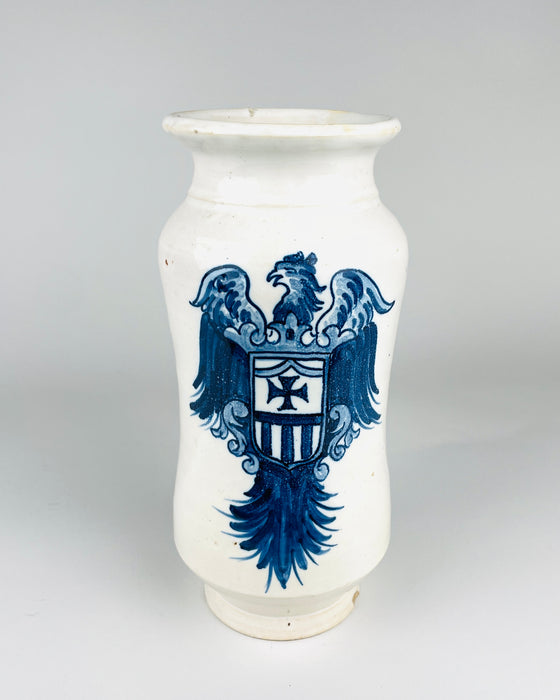 Large Antique Spanish Talavera albarello apothecary jar in glossy white glazed earthenware, decorated with a cobalt blue heraldic eagle carrying a shield with a cross and vertical stripes. The tall wheel thrown vessel has rounded shoulders, gently waisted sides, a flared rim and a circular foot. The decorative pharmacy jar is a distinctive example of Spanish blue and white pottery.