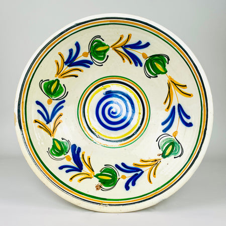 Antique Spanish lebrillo bowl in glazed earthenware, dating to the early 20th century and decorated in the Castilian remolino tradition. A cobalt blue spiral fills the centre while green vegetal forms, honey yellow branches and black accents rotate across the cream ground. The shallow flared basin works as ceramic wall art or a decorative bowl.