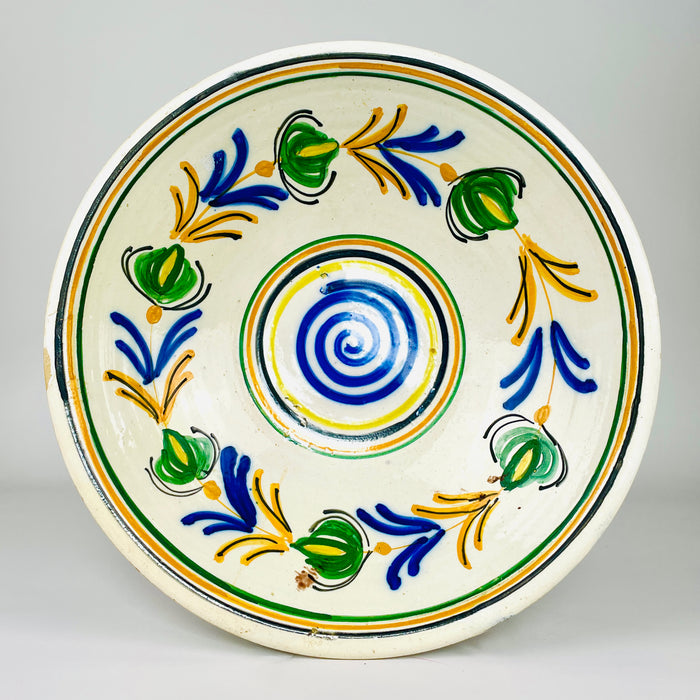 Antique Spanish lebrillo bowl in glazed earthenware, dating to the early 20th century and decorated in the Castilian remolino tradition. A cobalt blue spiral fills the centre while green vegetal forms, honey yellow branches and black accents rotate across the cream ground. The shallow flared basin works as ceramic wall art or a decorative bowl.
