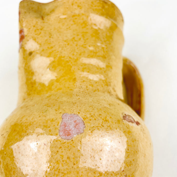 Nineteenth century Spanish pottery pitcher with a red clay body beneath a luminous mustard yellow glaze. The rounded vessel is wheel thrown and fitted with a curved handle, pinched pouring spout and low foot, while chips, glaze loss, crazing and rubbed edges record its utilitarian history as an everyday household jug.