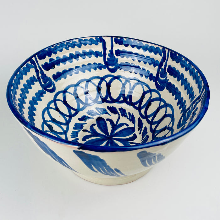 Large footed Fajalauza lebrillo bowl from Granada, hand painted by Cerámica Los Arrayanes in cobalt blue on cream glazed earthenware. Its deep flared form reveals a central flower, looping geometric border, wavy concentric bands and stylized plant motifs, giving the Spanish pottery bowl a sculptural presence for serving or Mediterranean decor.