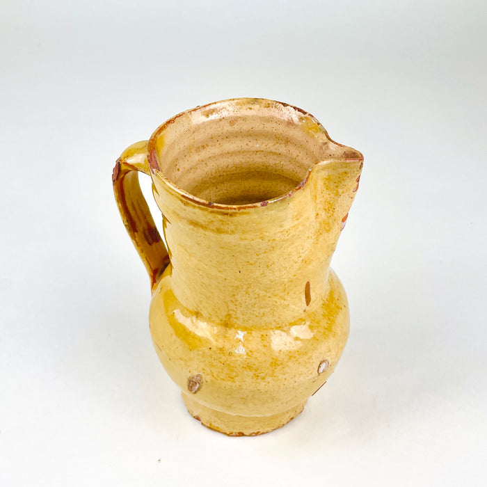 Small antique Spanish terracotta jug weighing 500 g, with a rounded belly and tall cylindrical neck balanced by a narrow circular foot. The yellow ceramic glaze shows natural tonal variation and substantial wear, while the pinched lip and sturdy handle identify the piece as a traditional liquid pitcher now suited to decorative farmhouse display.