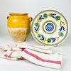 Spanish folk art lebrillo paired with a yellow earthenware vase and colorful striped cloths, highlighting the hand painted bowl as an early 20th century decorative object. Its central blue spiral, green floral wreath and concentric bands create a strong visual anchor for Mediterranean decor, a ceramic collection or a kitchen wall display.